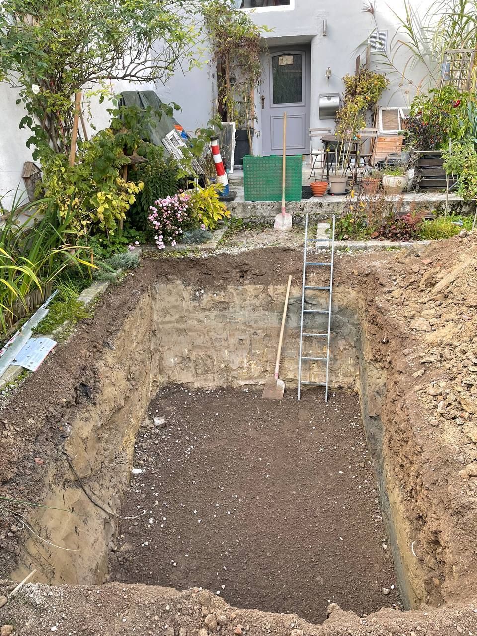 Ausgehobene Baugrube im Hinterhof mit Kiesboden, Leiter und weißer Hausterrasse im Hintergrund