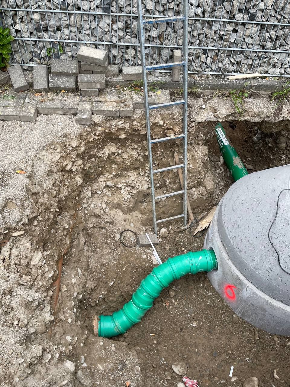 Eine Leiter in einem Erdgraben neben grünen Abflussrohren und einem grauen Versorgungstank in der Nähe einer Steinmauer.