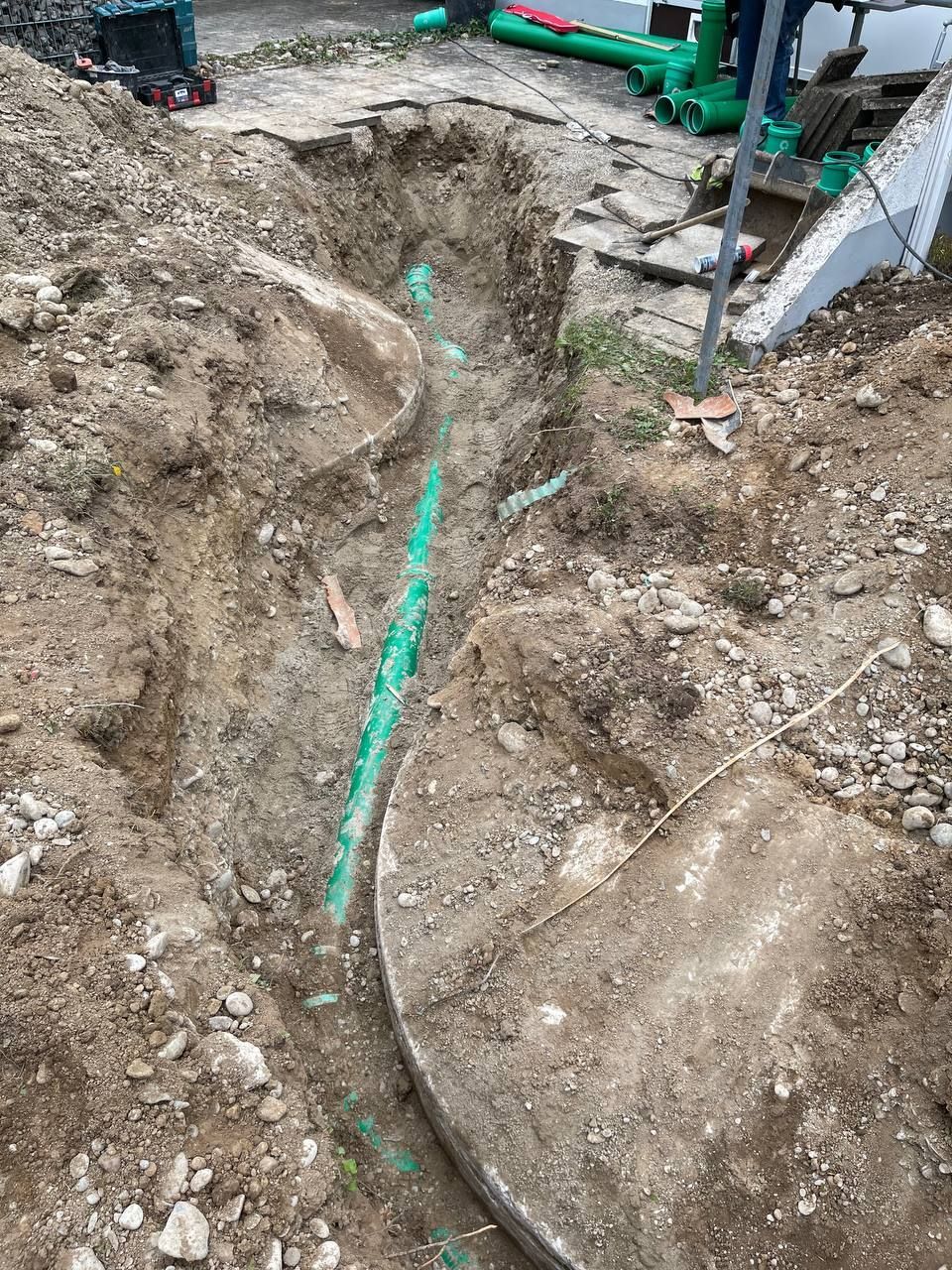 Aushubgraben in schlammigem Boden, durch den ein grünes Rohr verläuft, und Baumaschinen in der Nähe