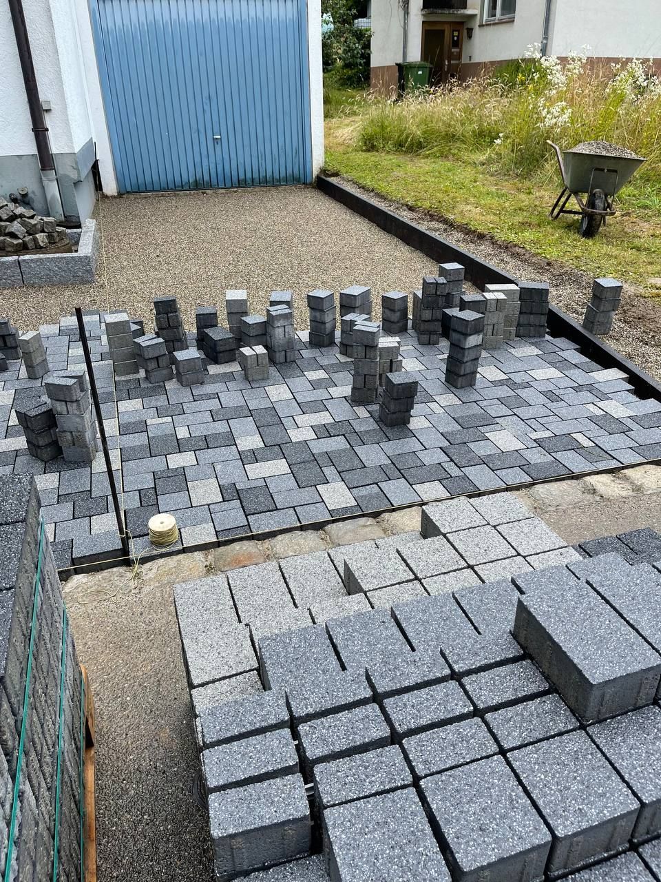 Außenterrasse mit gestapelten grauen Pflastersteinen und einem teilweise verlegten schachbrettartig gemusterten Steinweg in einem Gartenbereich
