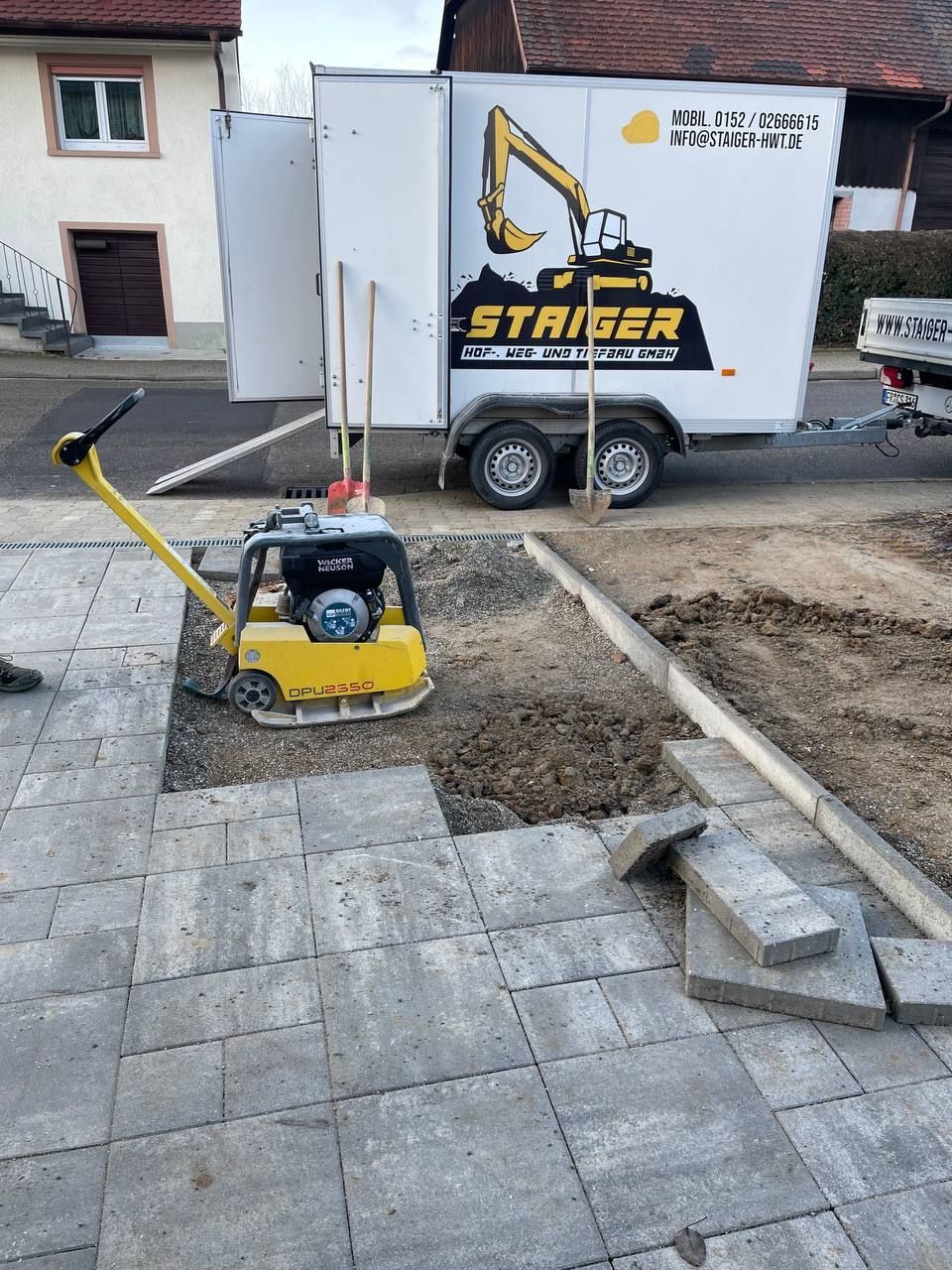 Baustelle mit einer gelben Rüttelplatte auf Pflastersteinen und einem STARKER-Geräteanhänger im Hintergrund