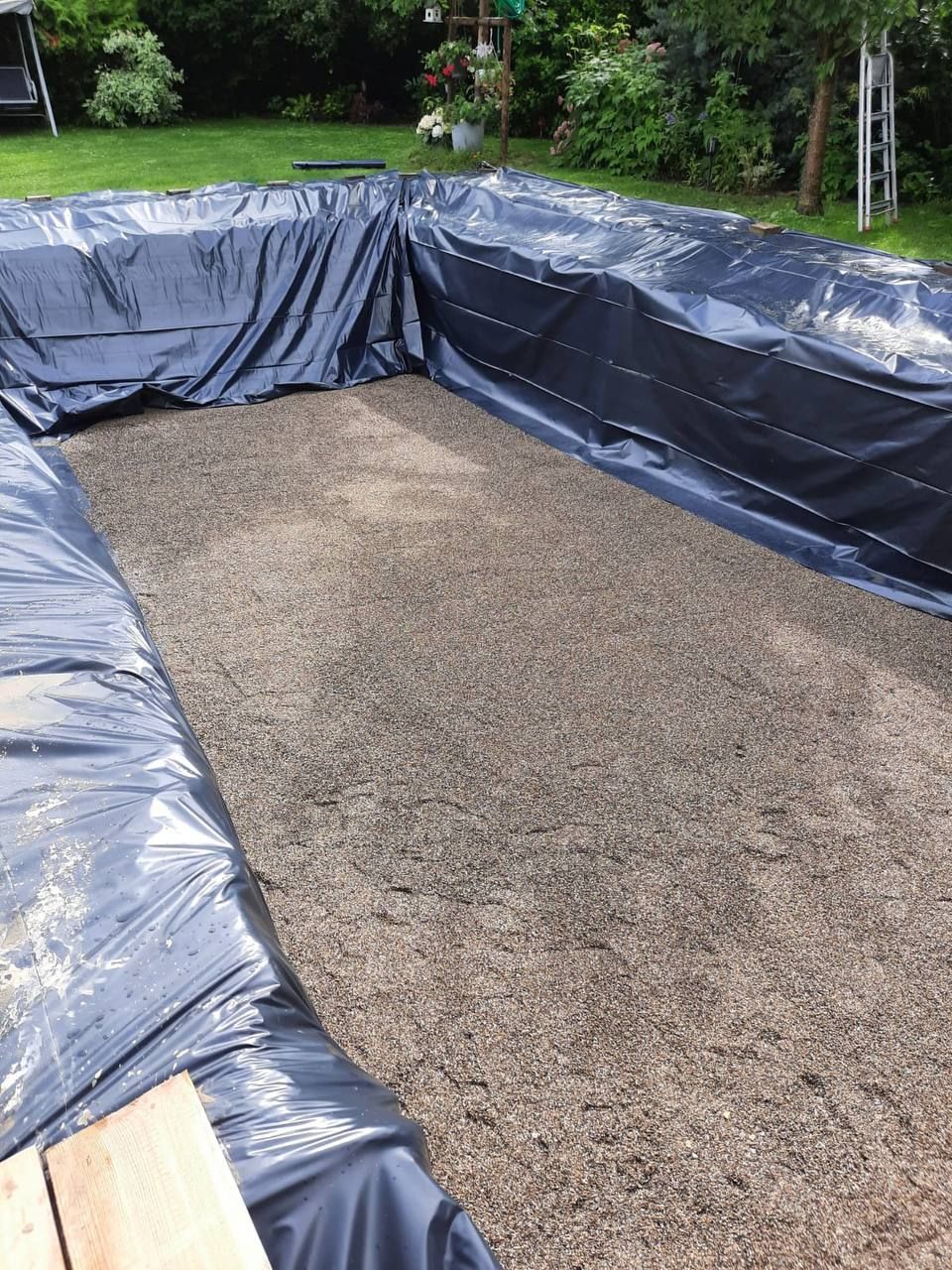 Mit Kies gefüllter Gartenteich, der mit schwarzer Plastikfolie ausgekleidet und von Gras und Holzplanken eingefasst ist