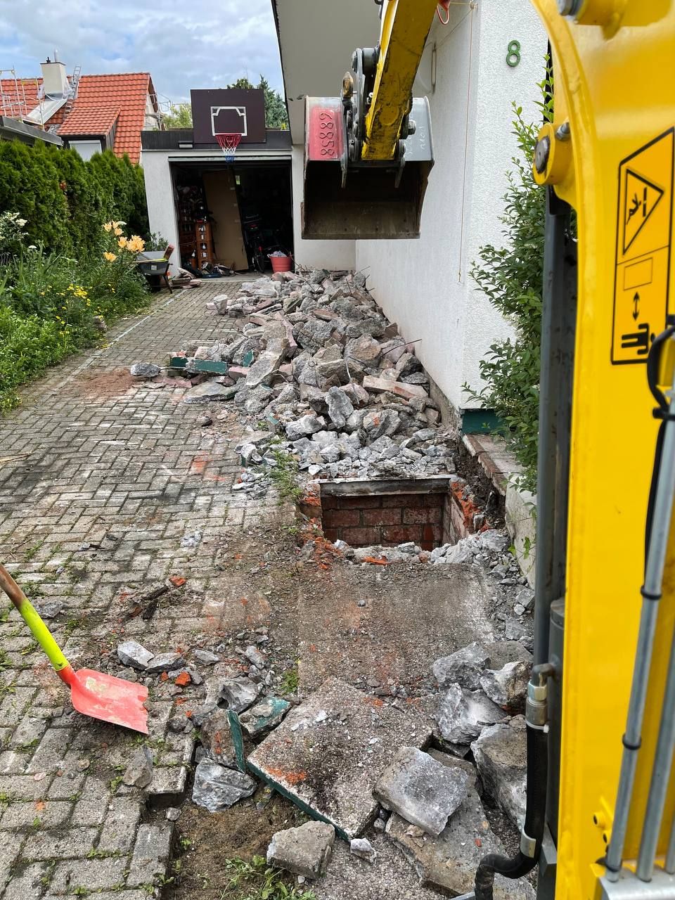 Ein Bagger räumt Schutt von einer gepflasterten Auffahrt neben einem weißen Haus und einer Garage.