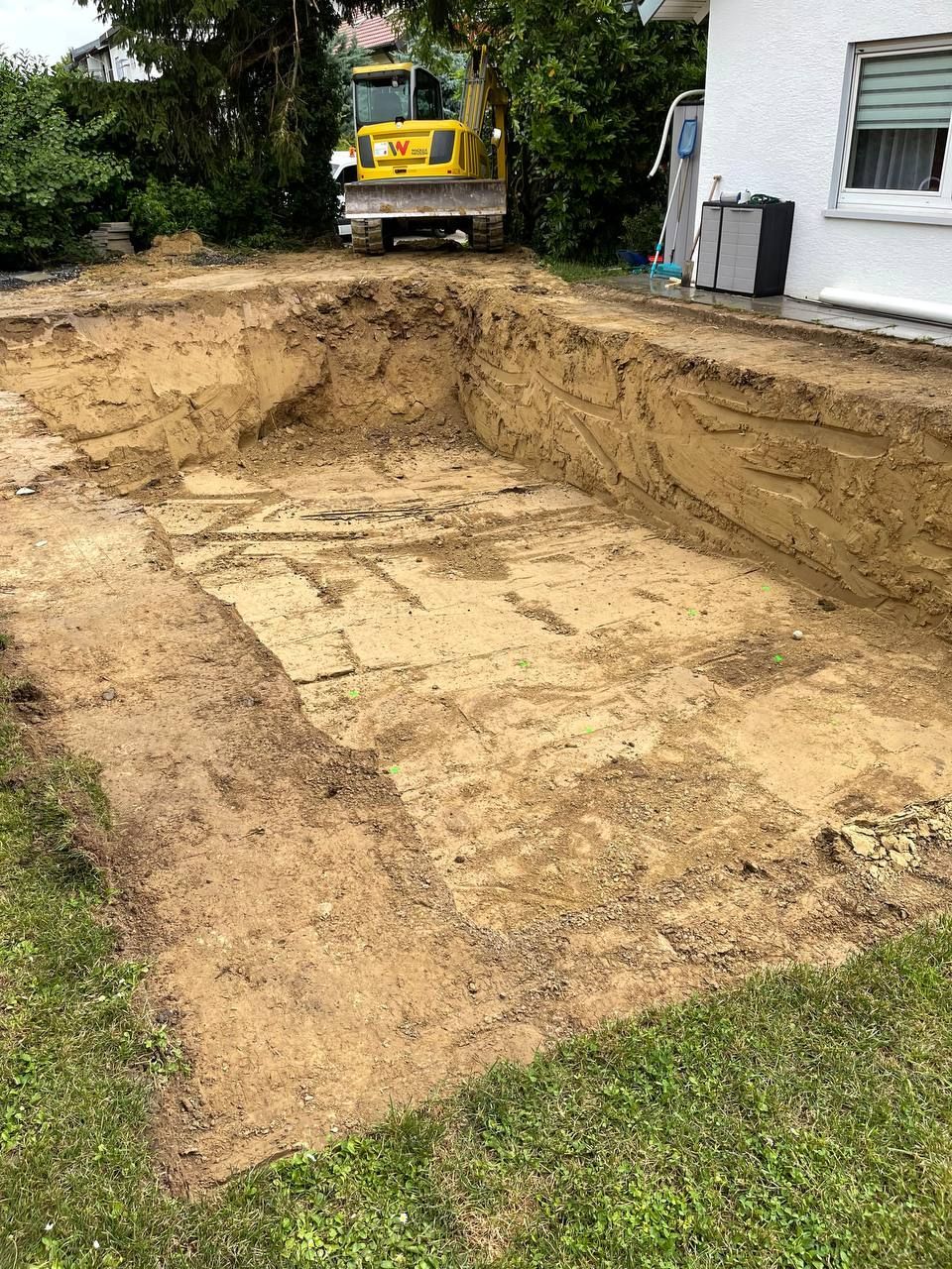 Ausgehobene Erdfläche neben einem Haus, im Hintergrund ein gelber Bagger, an den Rändern Gras.