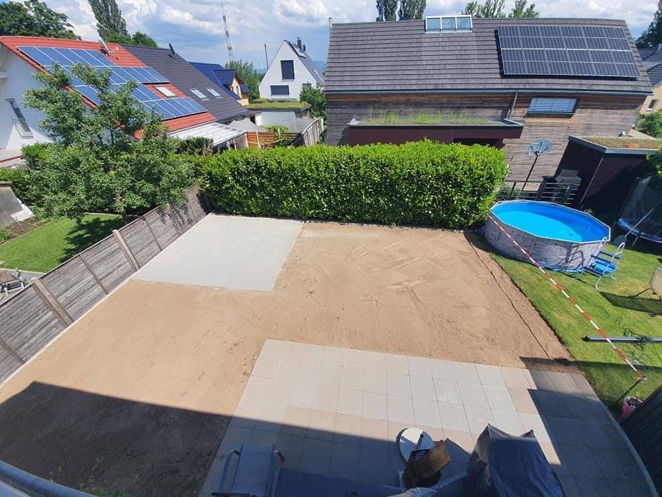 Hinterhofterrasse mit unfertigem Betonboden, Heckenbegrenzung und einem blauen Trampolin neben benachbarten Häusern und Solardächern.