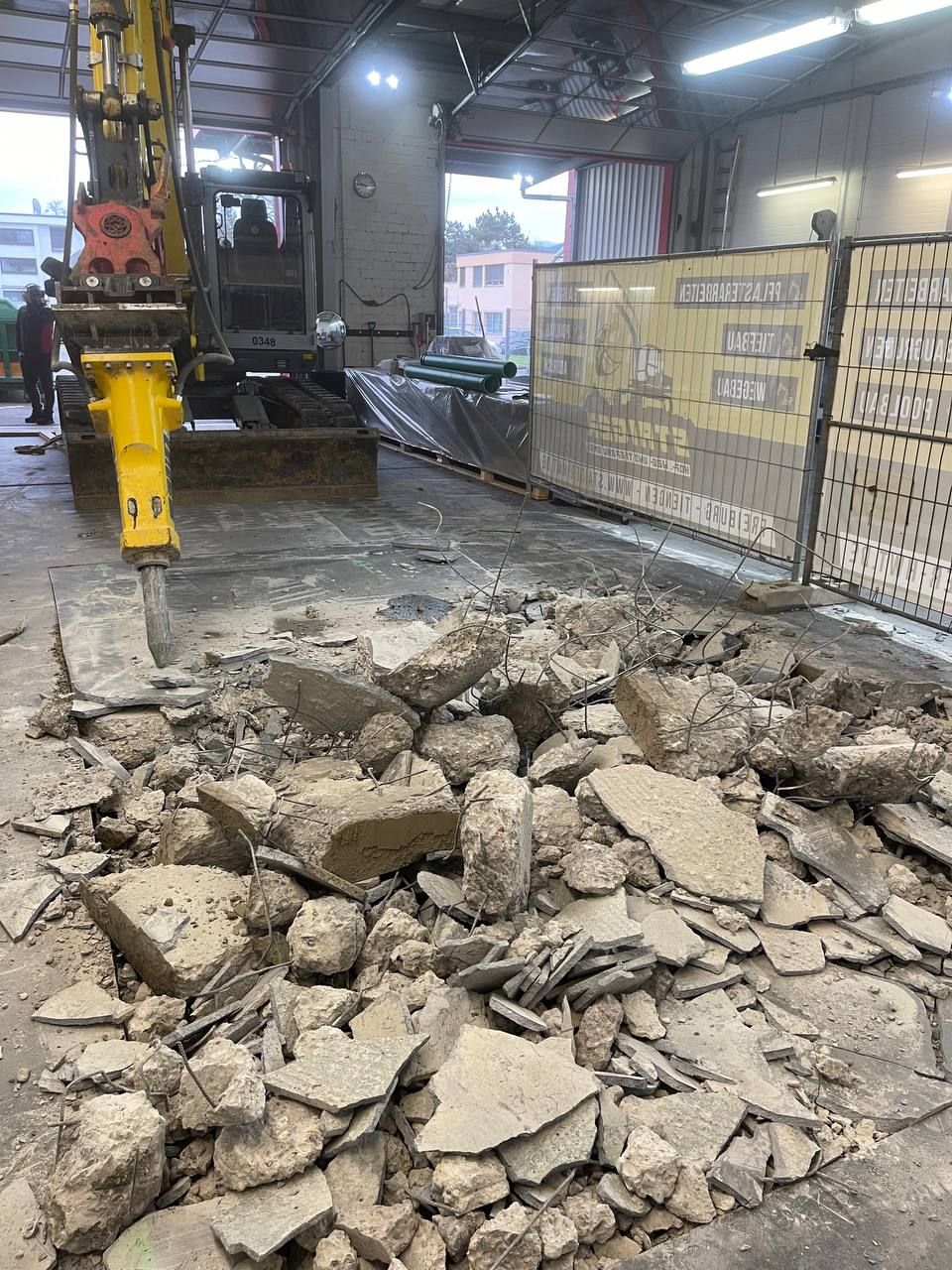 Ein Bagger zerkleinert Betonschutt in einer Industriehalle; im Vordergrund ist der Schutt aufgehäuft.