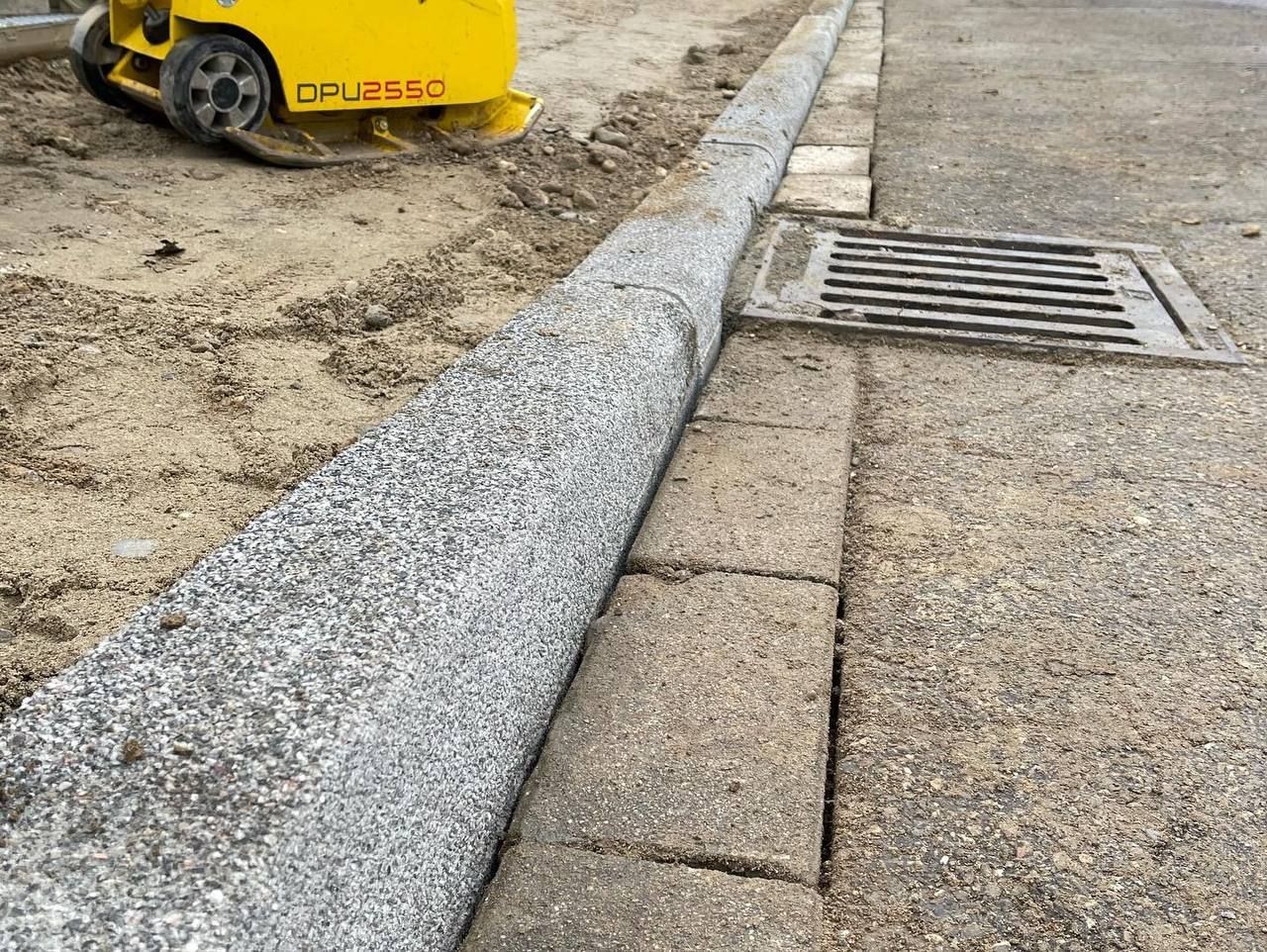 Straßenbauarbeiten mit Bordsteinkante, frischem Kies, Pflastersteinen und einem Regenwasserablaufgitter; Baumaschinen