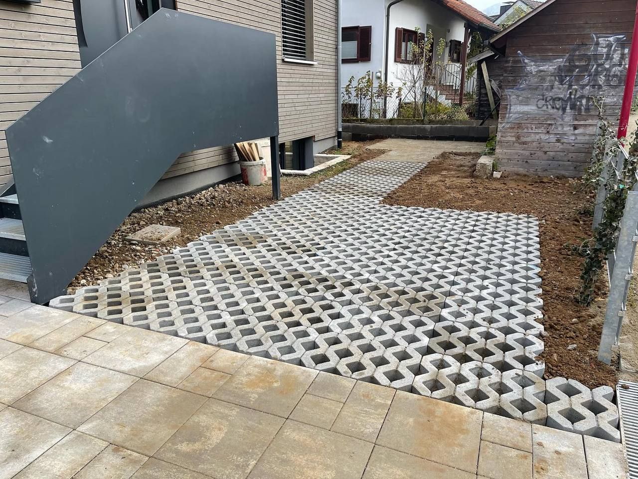 Unbebautes Grundstück mit weißen Gitterpflastersteinen, Kies und einer großen dunklen Tafel neben einem Haus