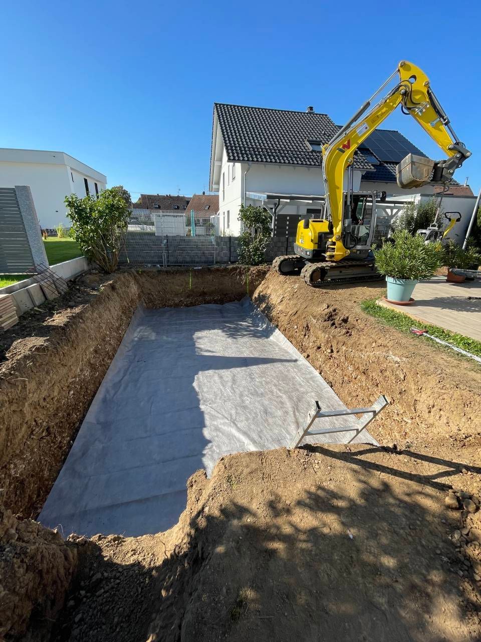 Ein Minibagger hebt im Garten neben einem Haus einen Graben aus, der mit Kies bedeckt ist und aus Erdhaufen besteht.
