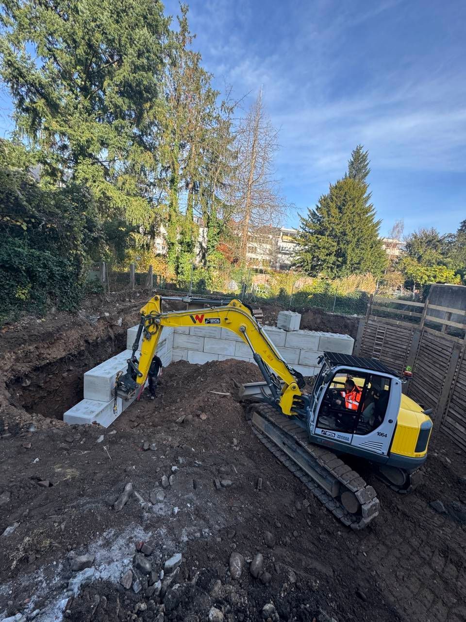 Ein gelber Bagger gräbt Erde neben einem Betonfundament in einem bewaldeten Hinterhofgarten aus.