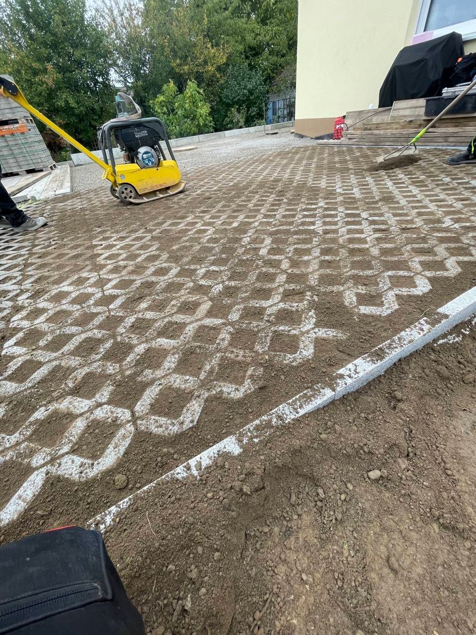 Baustelle mit einer gelben Rüttelplatte auf mit weißen Geogitterlinien überzogenem Kies.