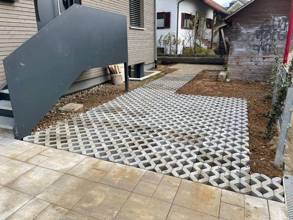 Kiesparkplatz mit weißen Kunststoffgitterplatten neben einem Haus und gepflastertem Gehweg.