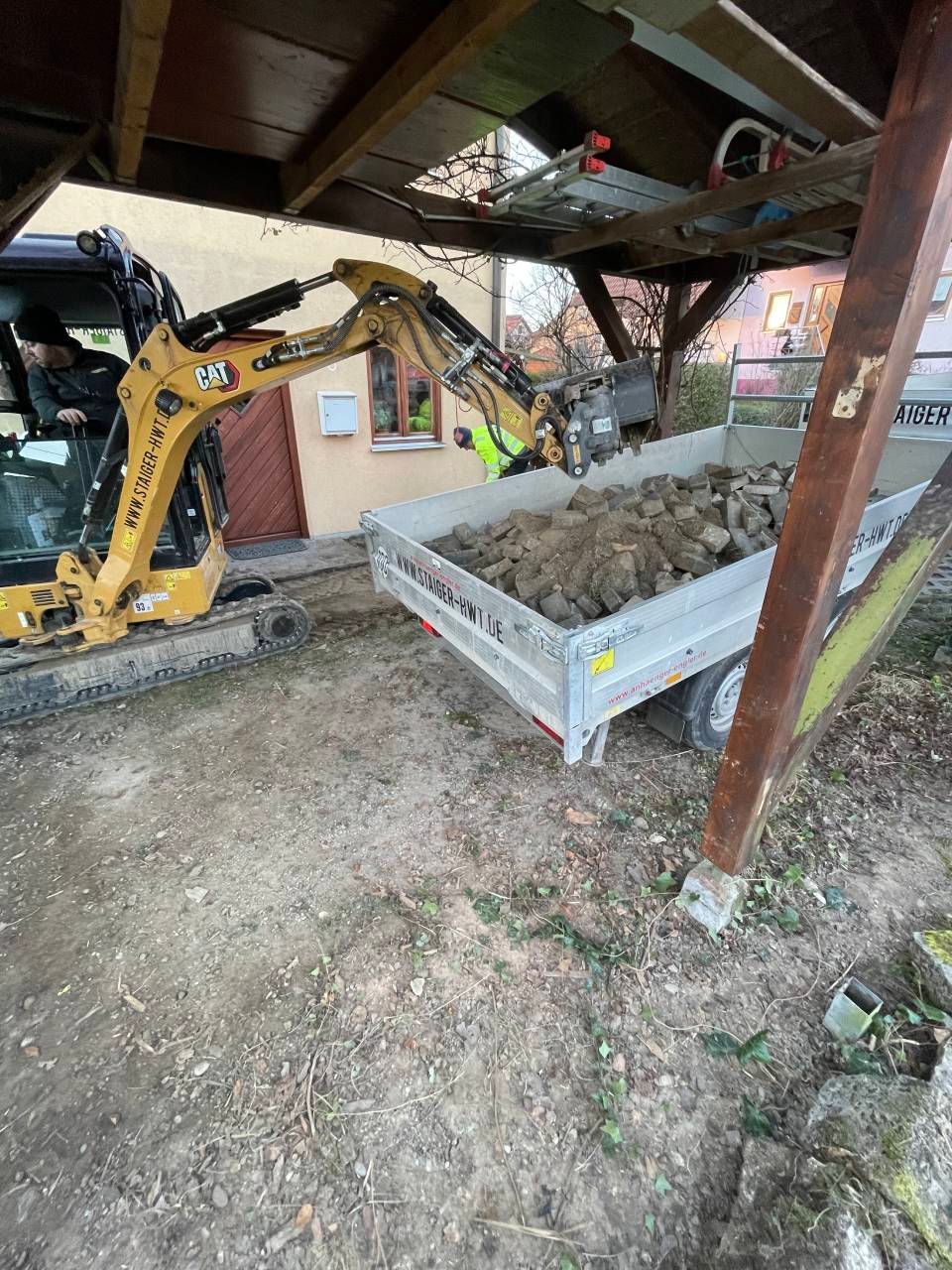 Ein gelber Minibagger verlädt Erde in einen Anhänger unter einem Schuppen.