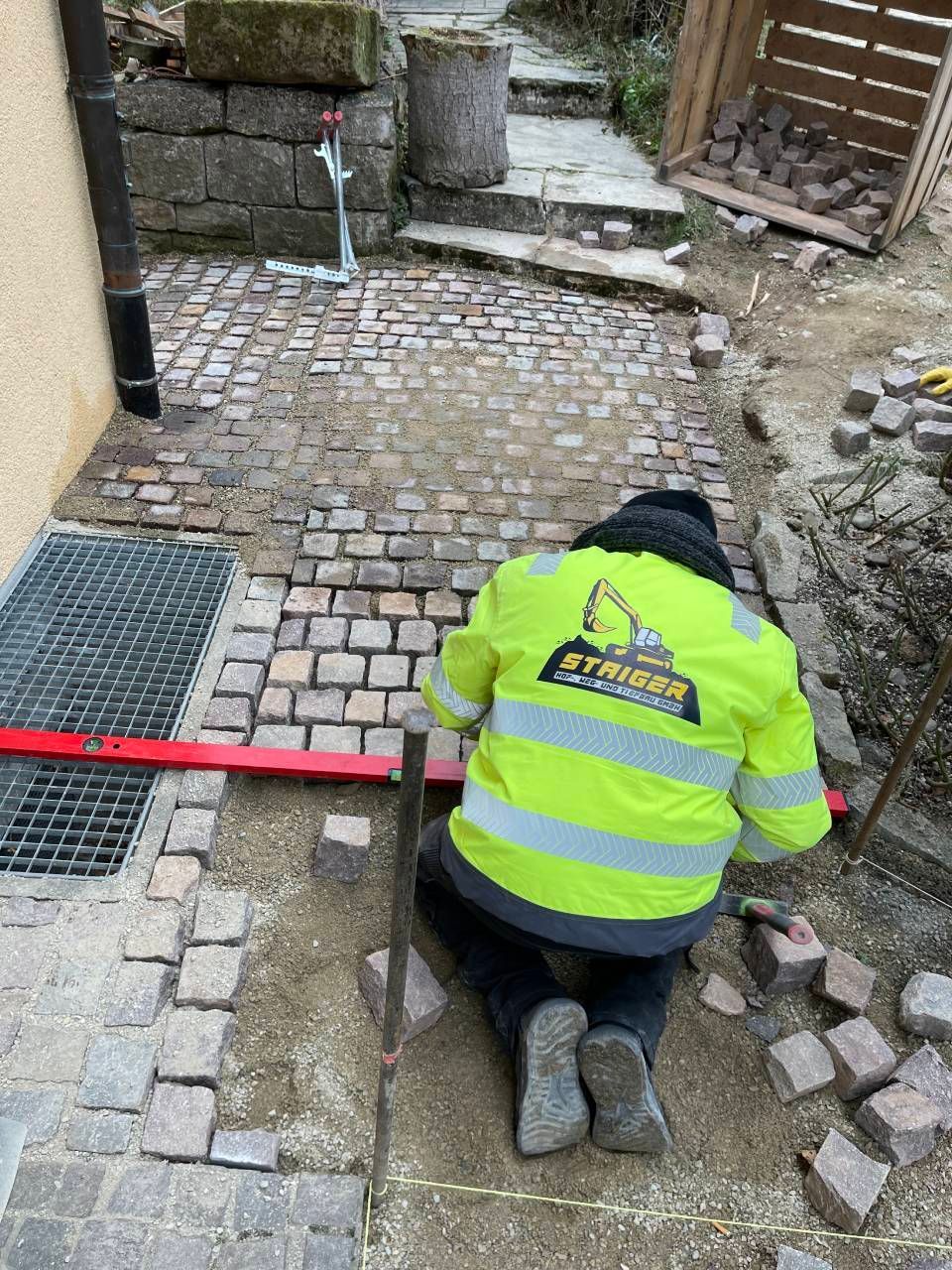 Ein Arbeiter in einer neongelben Jacke kniet auf Kopfsteinpflaster neben einem roten Absperrband und repariert den Gehweg.