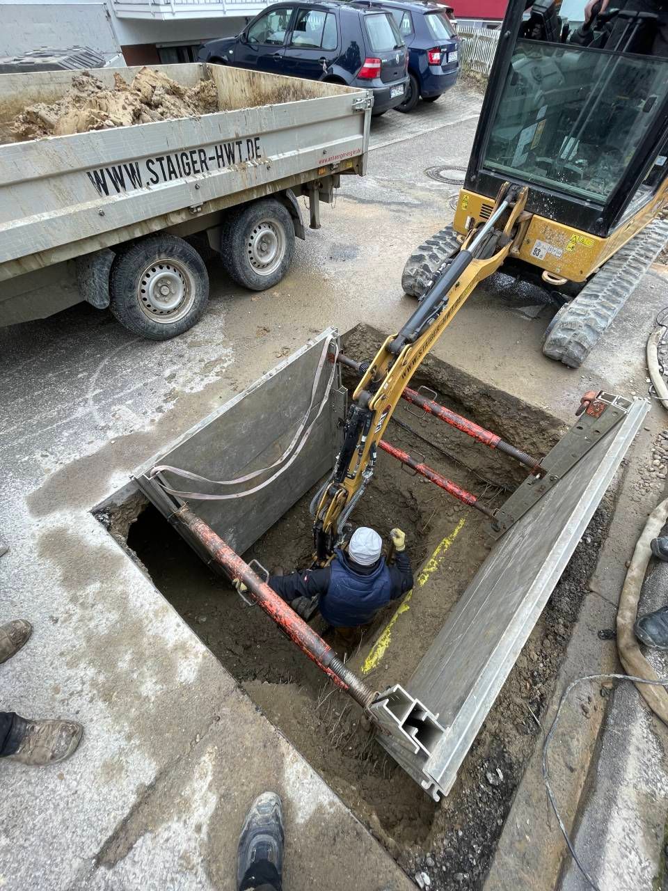 Ein Arbeiter steht in einem Graben neben einem Bagger und einem Muldenkipper auf einer Straßenbaustelle.