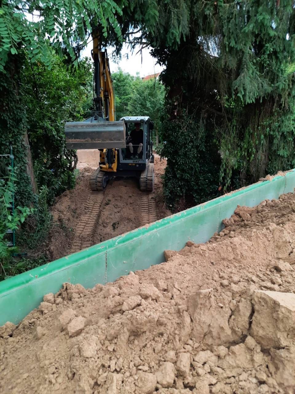 Bulldozer an einem Erdhang neben einer grünen Stützmauer, umgeben von Bäumen