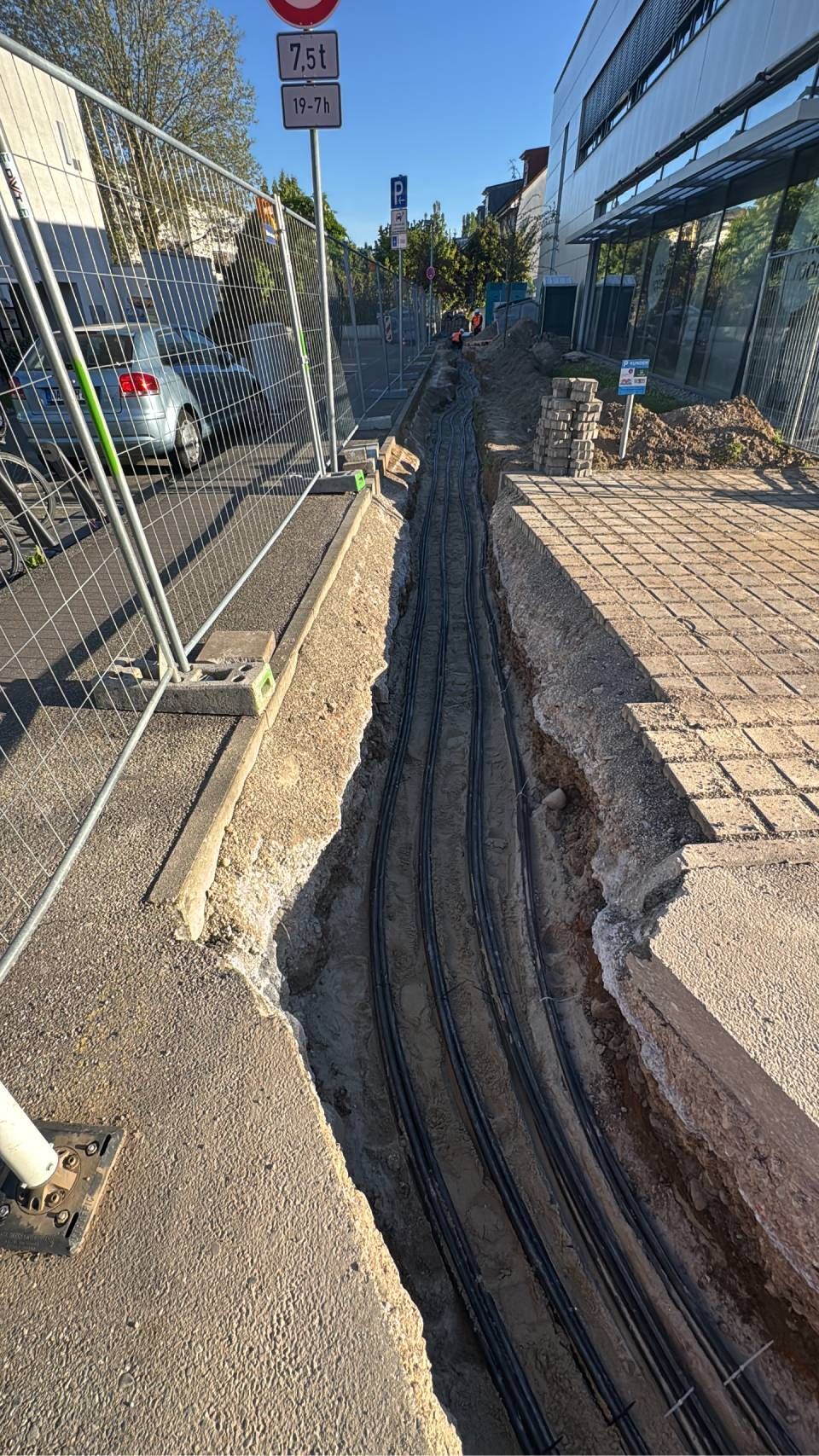 Ein schmaler Gehweggraben neben einem Maschendrahtzaun und geparkten Autos auf einer sonnigen Straße