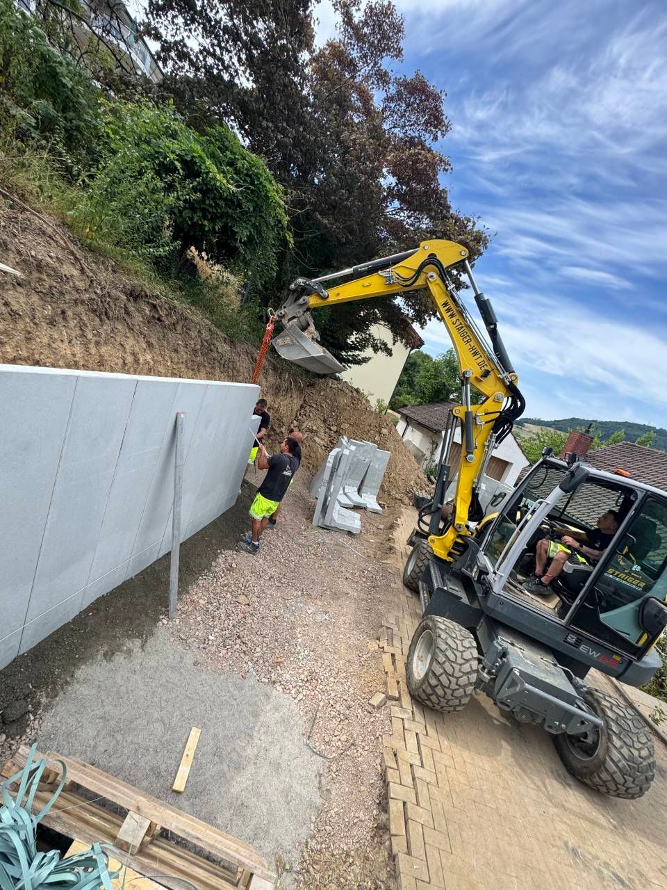 Ein Bagger hebt an einer Baustelle am Straßenrand eine Betonwandplatte an; Arbeiter befinden sich in der Nähe.