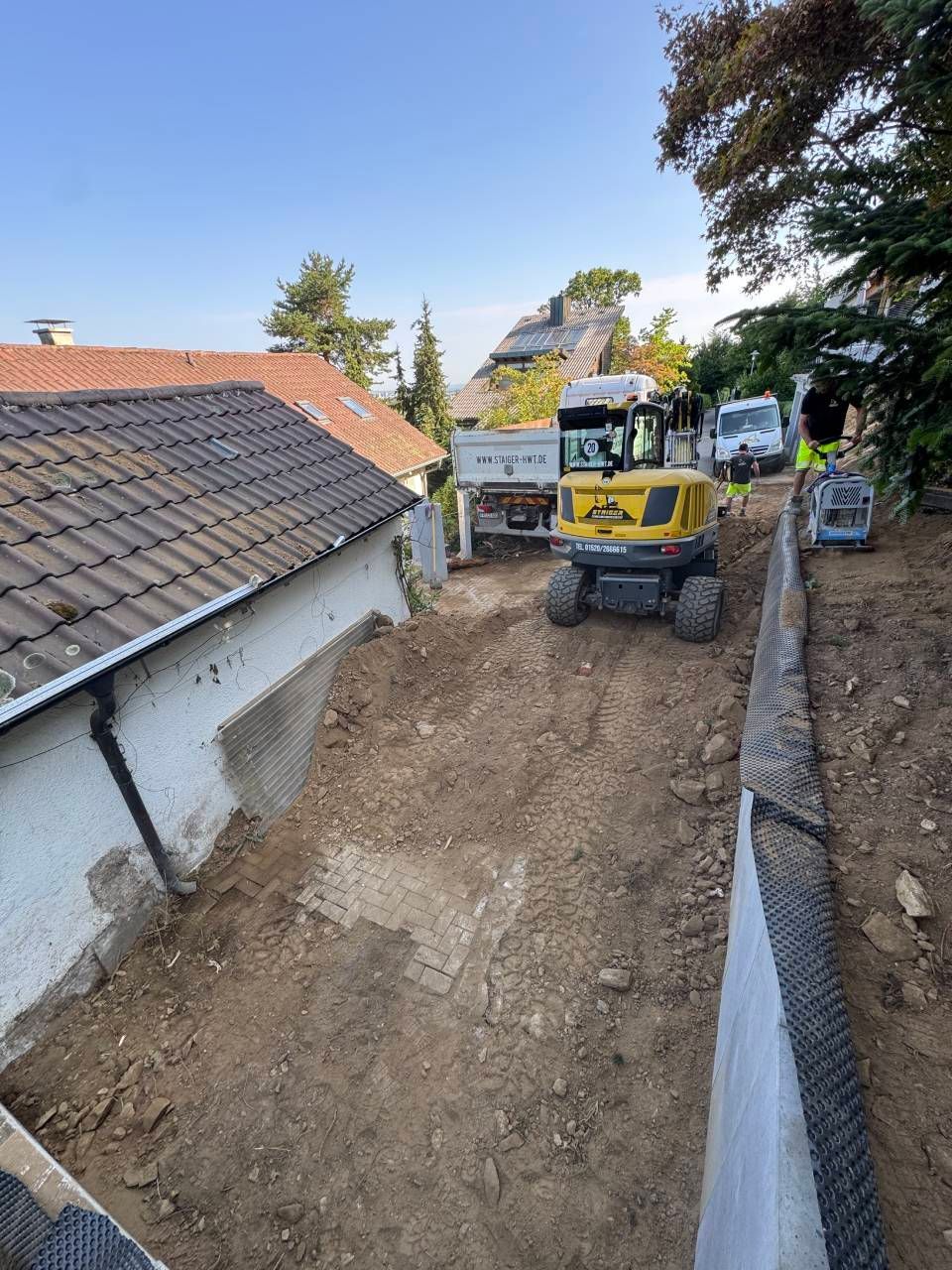 Baumaschinen heben einen schmalen Erdgraben neben einem Haus und einer Stützmauer aus.