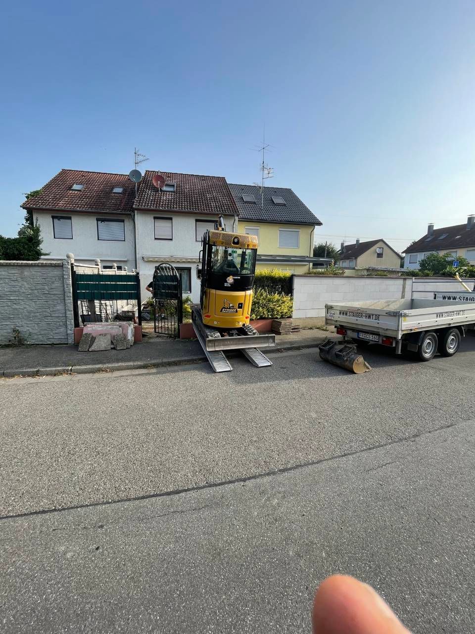 Vor einem Haus in einer ruhigen Straße parkten Baumaschinen, darunter ein kleiner gelber Bagger mit Anhänger.