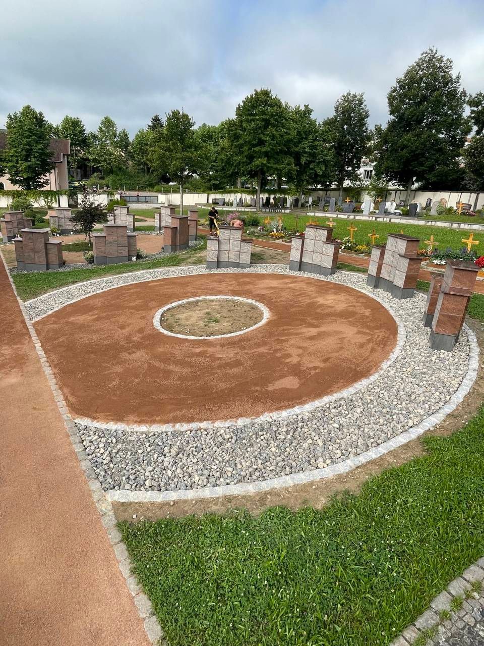 Runder Gedenkplatz aus Ziegelsteinen mit gravierten Steinsäulen, Kiesrand und Bäumen unter einem bewölkten Himmel