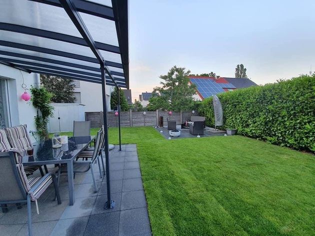 Terrasse mit überdachten Sitzgelegenheiten, gepflastertem Weg und grünem Rasen neben einer Hecke und einem kleinen Gartenhaus