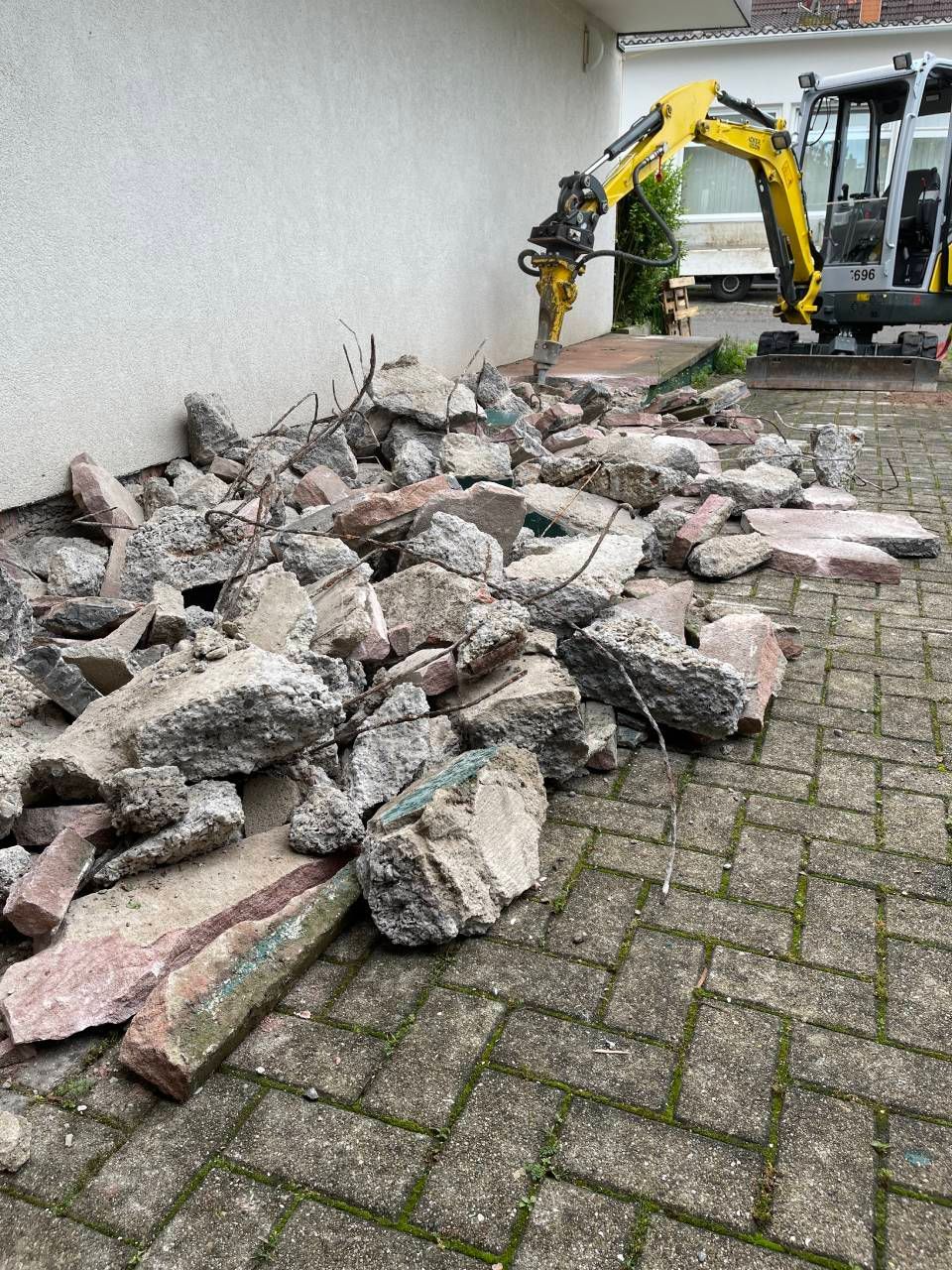 Ein Haufen zerbrochener Betontrümmer liegt neben einem gelben Bagger auf einer gepflasterten Auffahrt.