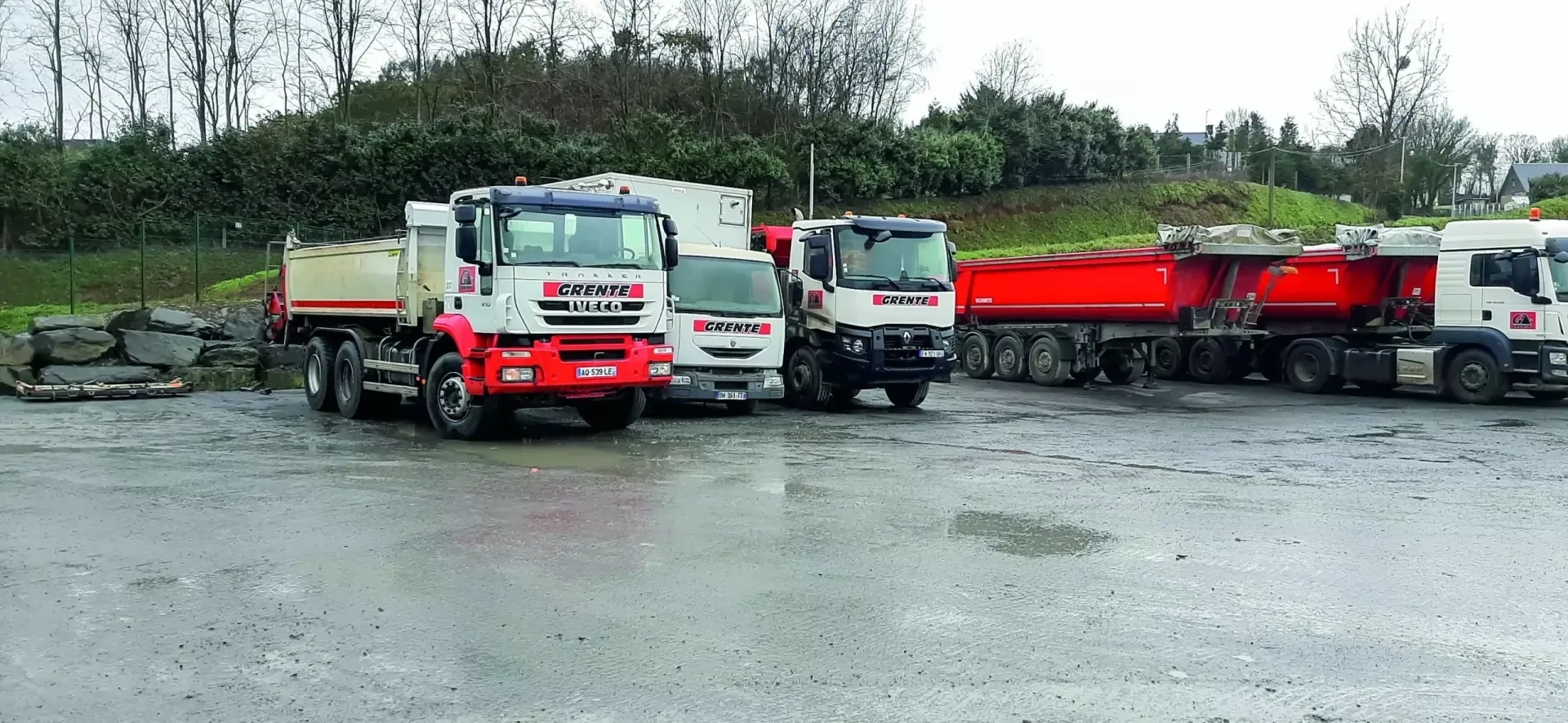 Plusieurs camions à benne basculante stationnés sur un terrain humide et gris avec un arrière-plan de buissons et d'arbres.
