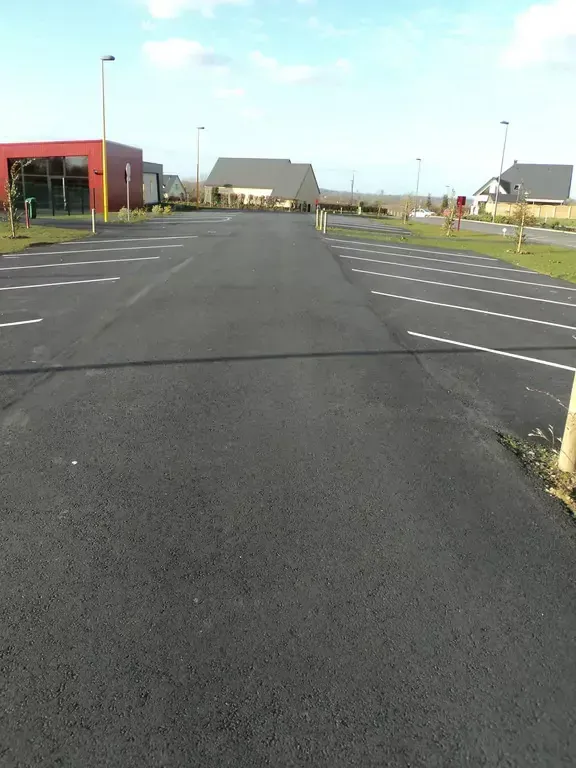 Parking asphalté vide avec des lignes de stationnement blanches, des maisons en arrière-plan et un bâtiment rouge sur la gauche.