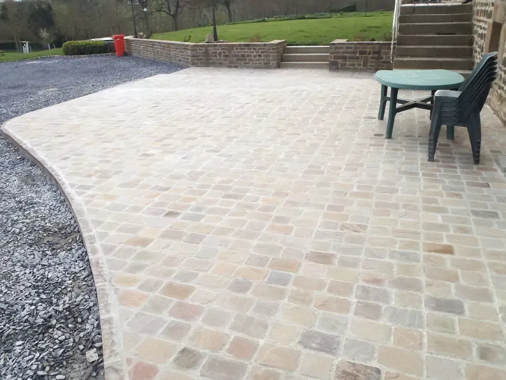Patio pavé avec un bord incurvé, zone de gravier et mobilier d'extérieur à côté d'un bâtiment et de marches, pelouse verte en arrière-plan.