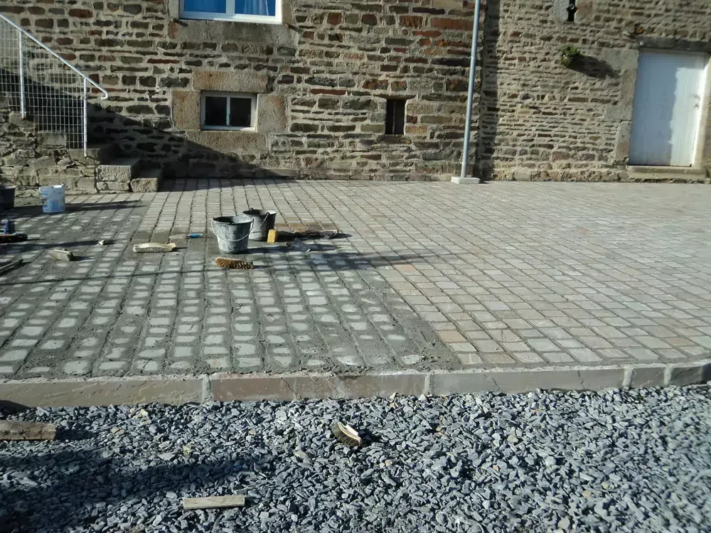 Patio extérieur en briques partiellement construit avec un bâtiment en pierre en arrière-plan.