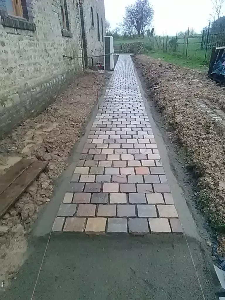 Construction d'un chemin en briques à côté d'un bâtiment en pierre. Briques posées dans du mortier, entourées de terre.