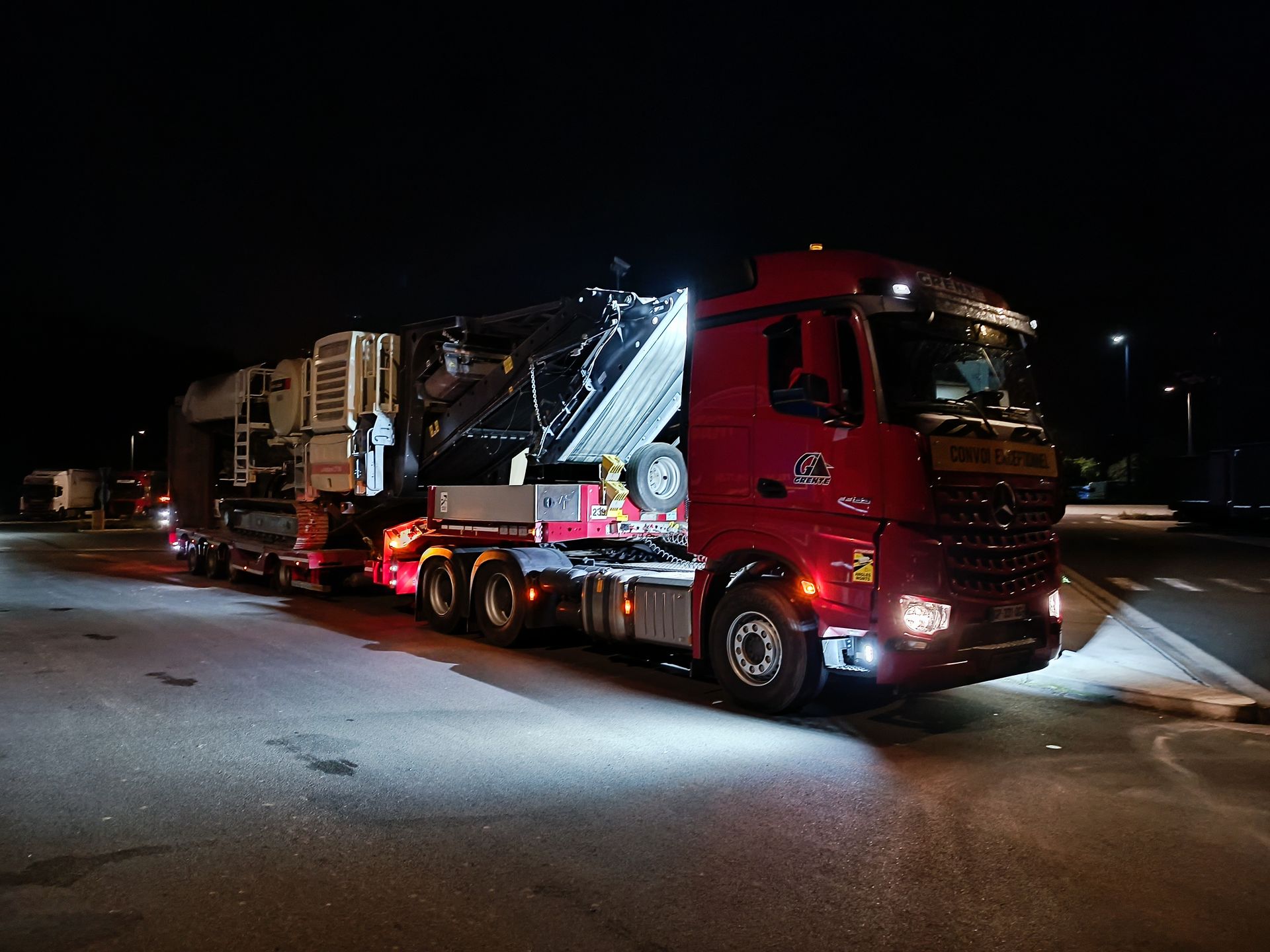 Un semi-remorque rouge transporte de grosses machines de nuit ; des phares illuminent le sol.
