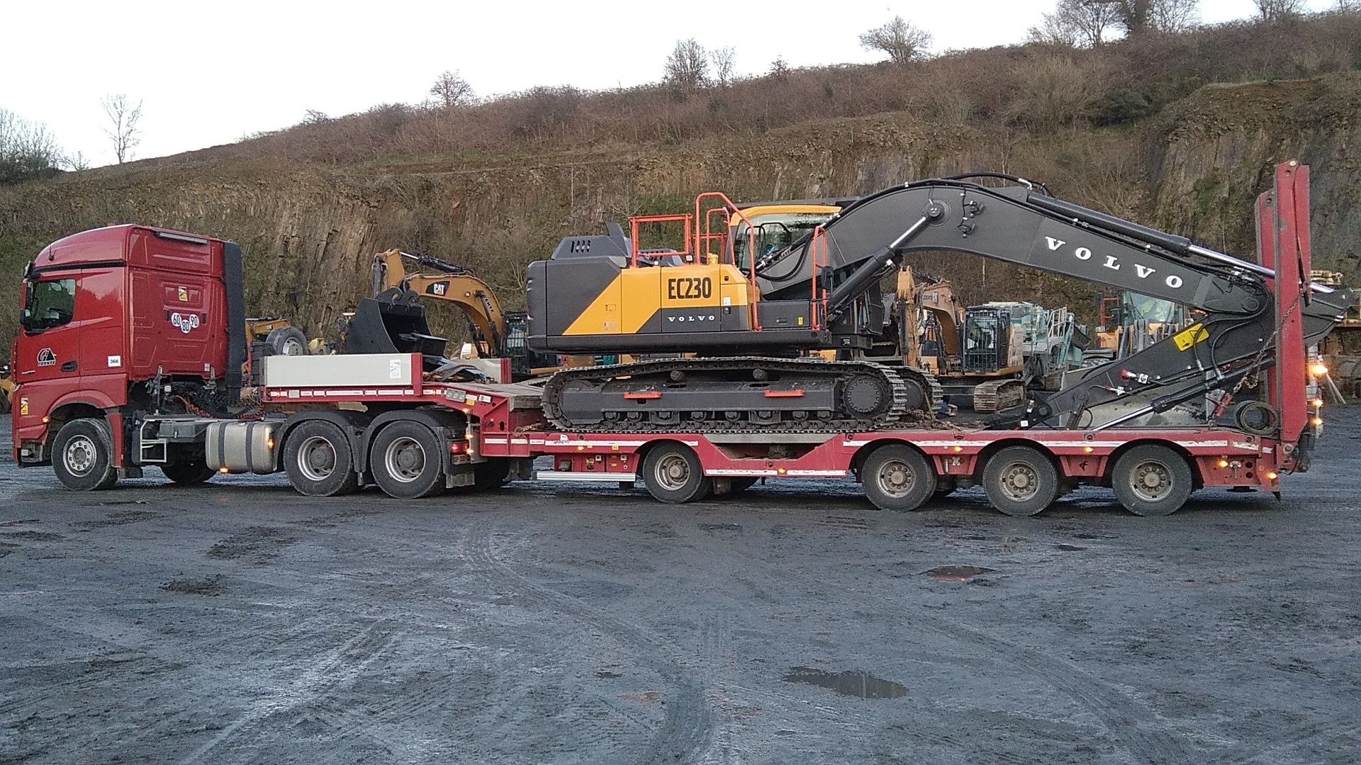 Un semi-remorque rouge transporte des engins lourds, dont une pelle mécanique Volvo, sur une remorque à plateau dans une carrière.