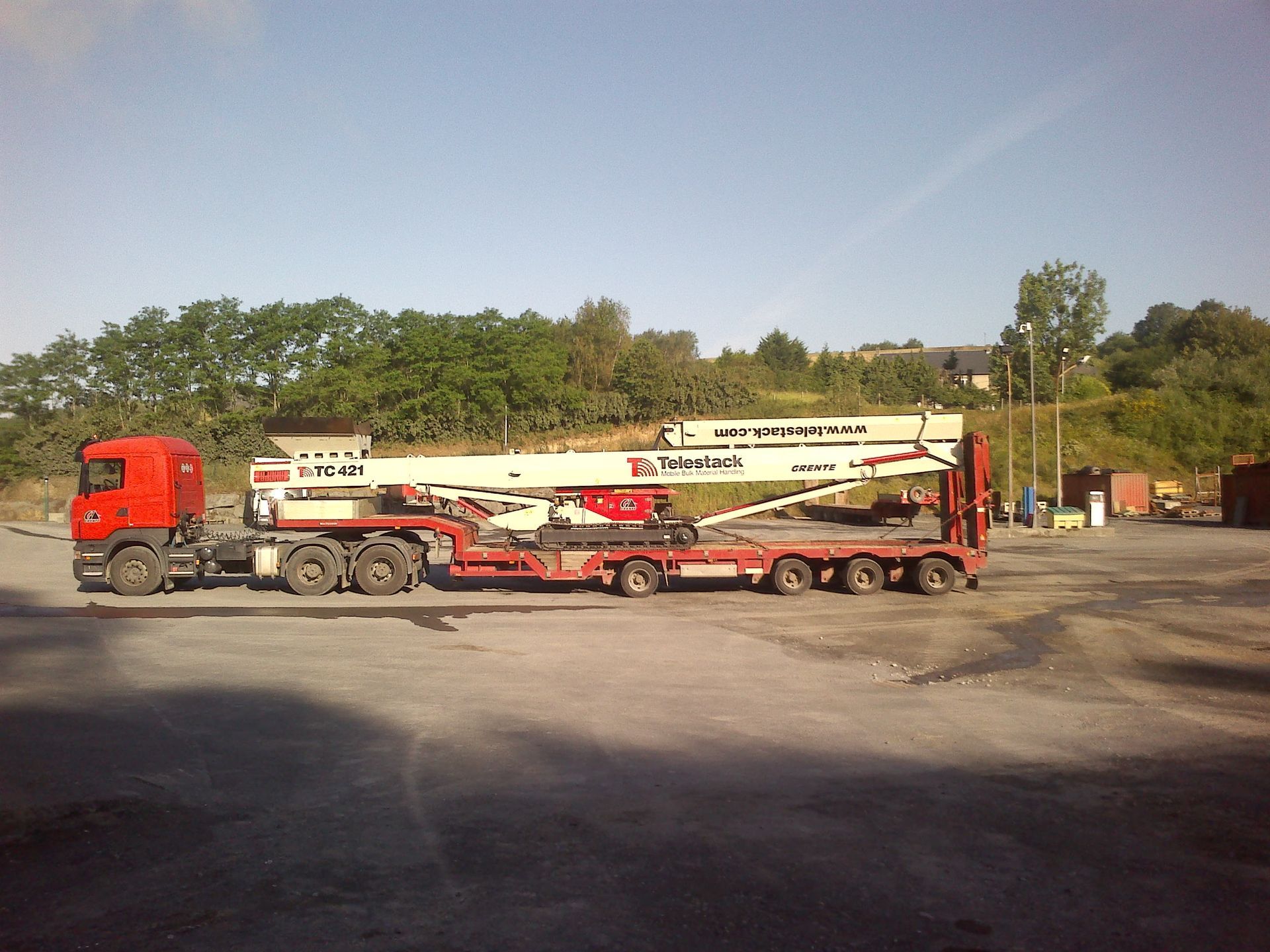 Un camion rouge tire une grosse machine de chantier blanche sur une remorque à plateau, sur un terrain gravillonné.