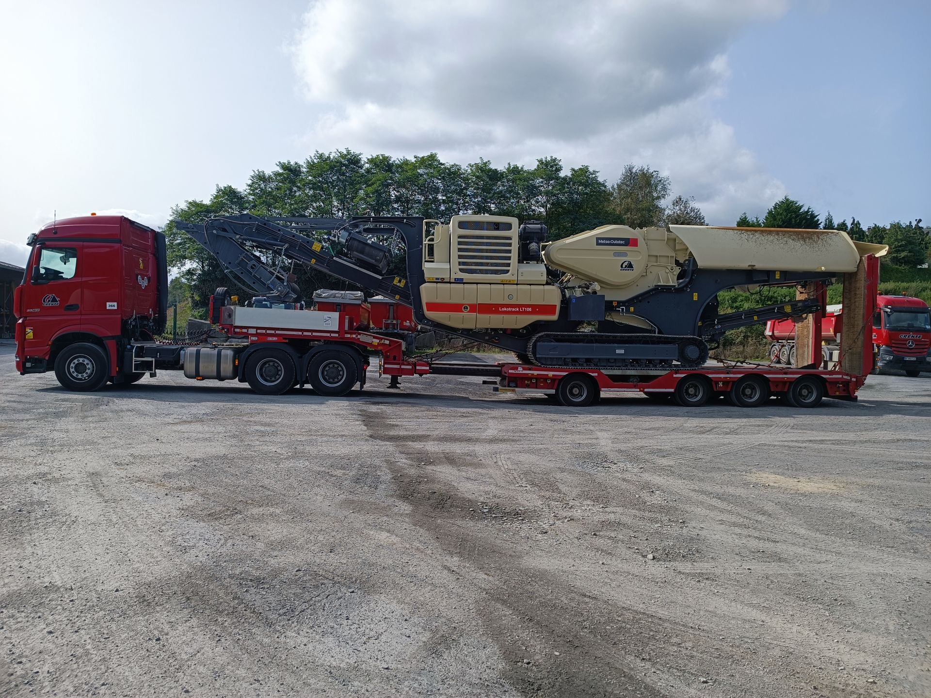 Un semi-remorque rouge transportant de grosses machines beiges sur une remorque surbaissée est stationné sur du gravier.