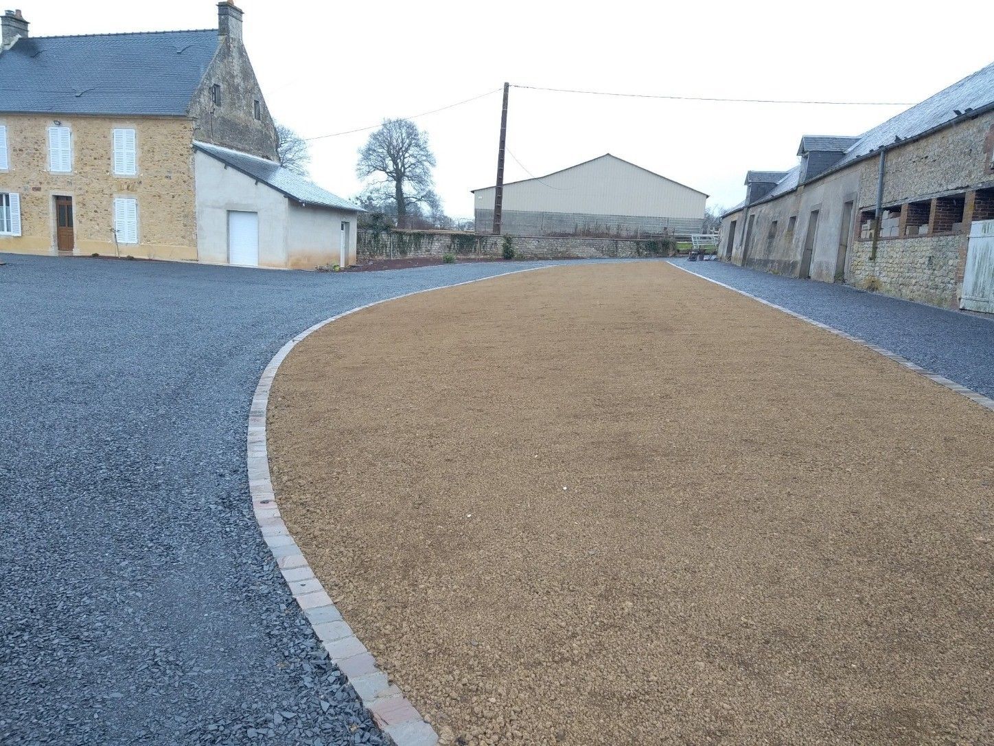 Allée de gravier avec une partie beige bordée de briques, devant des bâtiments en pierre et un ciel nuageux.