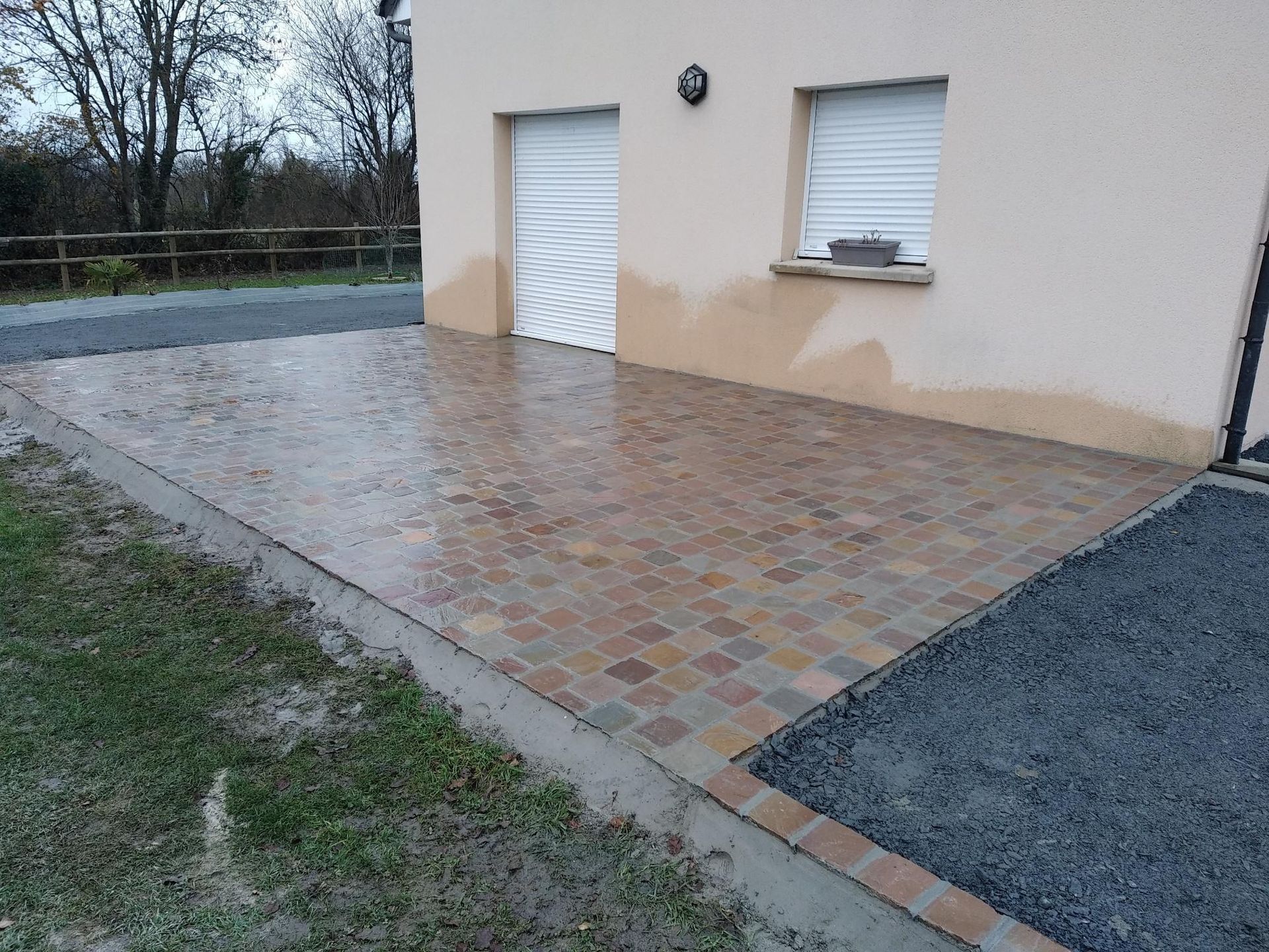 Terrasse pavée de dalles carrées jouxtant une maison dont la porte et la fenêtre blanches sont fermées.
