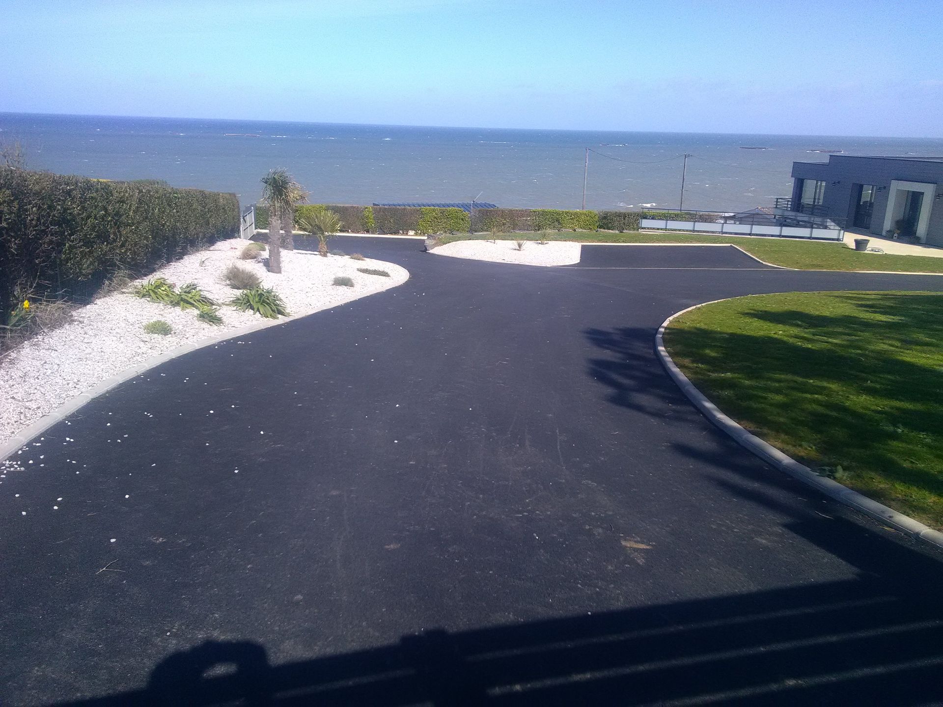 Allée asphaltée bordée de pierres blanches, jouxtant une pelouse. Océan en arrière-plan.