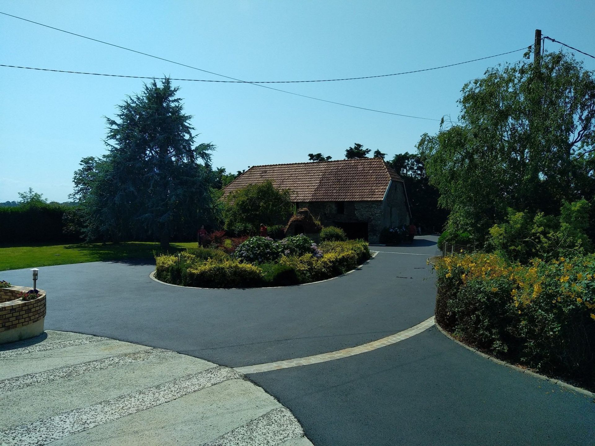 Allée asphaltée menant à une maison rustique en pierre entourée de verdure sous un ciel bleu.