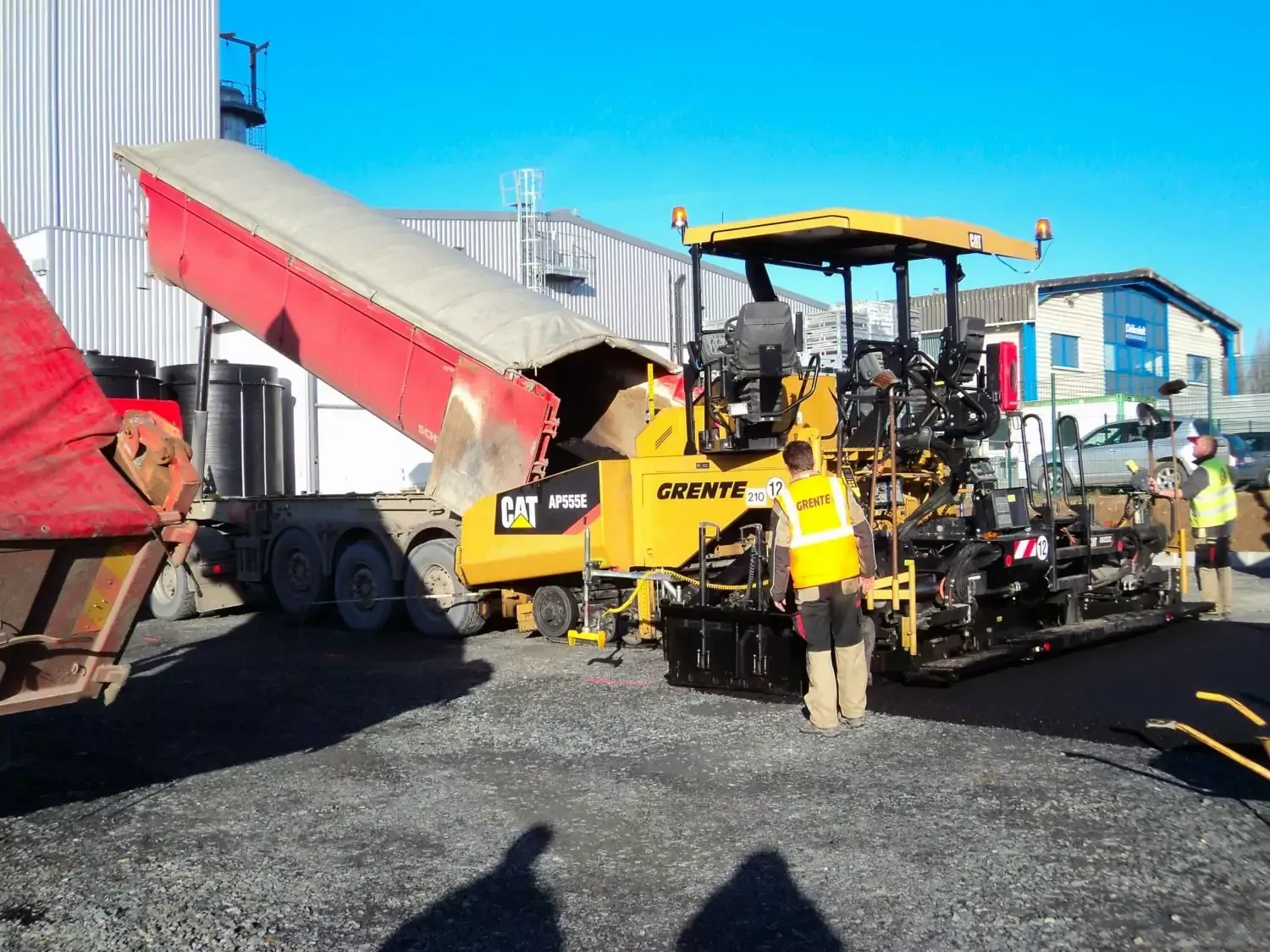 Une machine à paver l'asphalte jaune reçoit l'asphalte d'un camion à benne basculante rouge, des ouvriers portant des gilets de haute visibilité sont sur place.