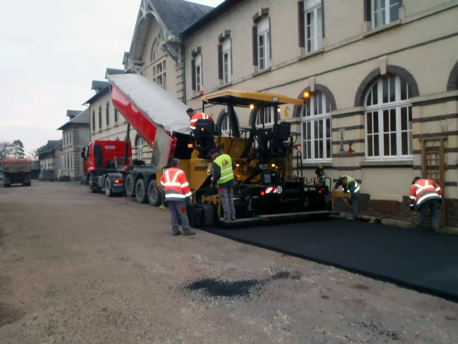 Travaux de pavage de route en cours près d'un bâtiment, l'asphalte est posé par une machine, les ouvriers sont en gilets.