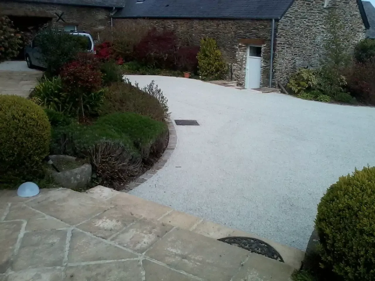 Allée en gravier blanc, bordée de haies basses et de marches en pierre menant à un bâtiment.