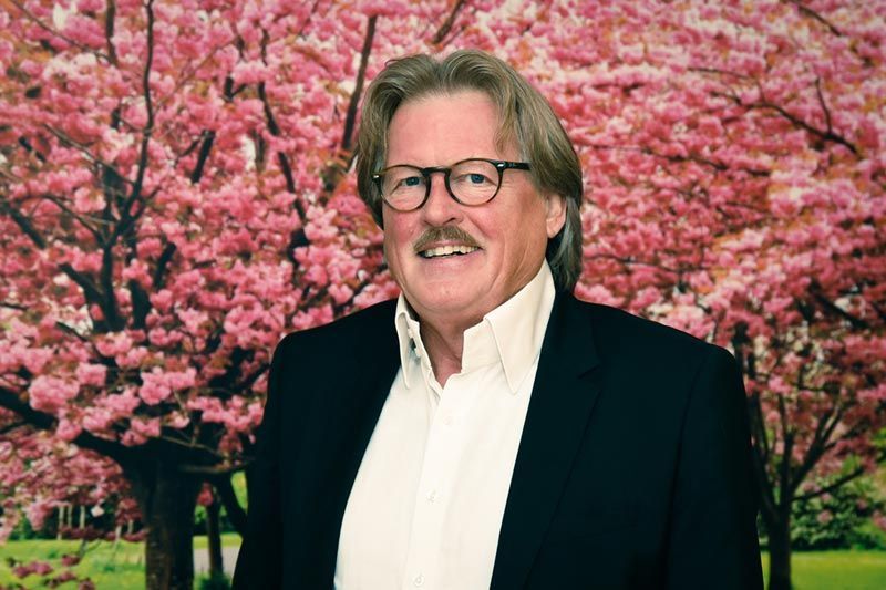 Ein Mann mit Brille und Anzug steht vor einem Kirschblütenbaum.