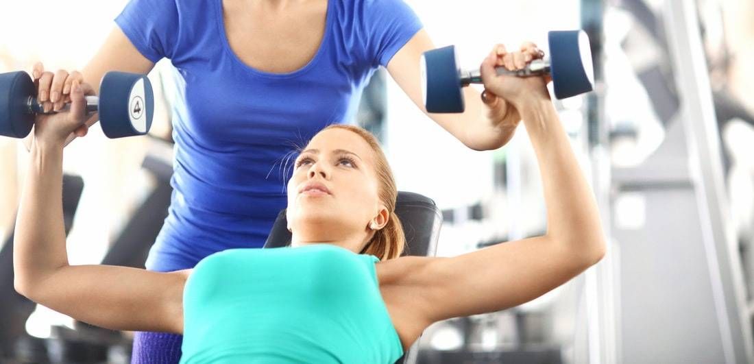 Ein Fitnesstrainer unterstützt eine Person bei der Ausführung einer Kurzhantel-Brustpresse auf einer Hantelbank in einem Fitnessstudio.