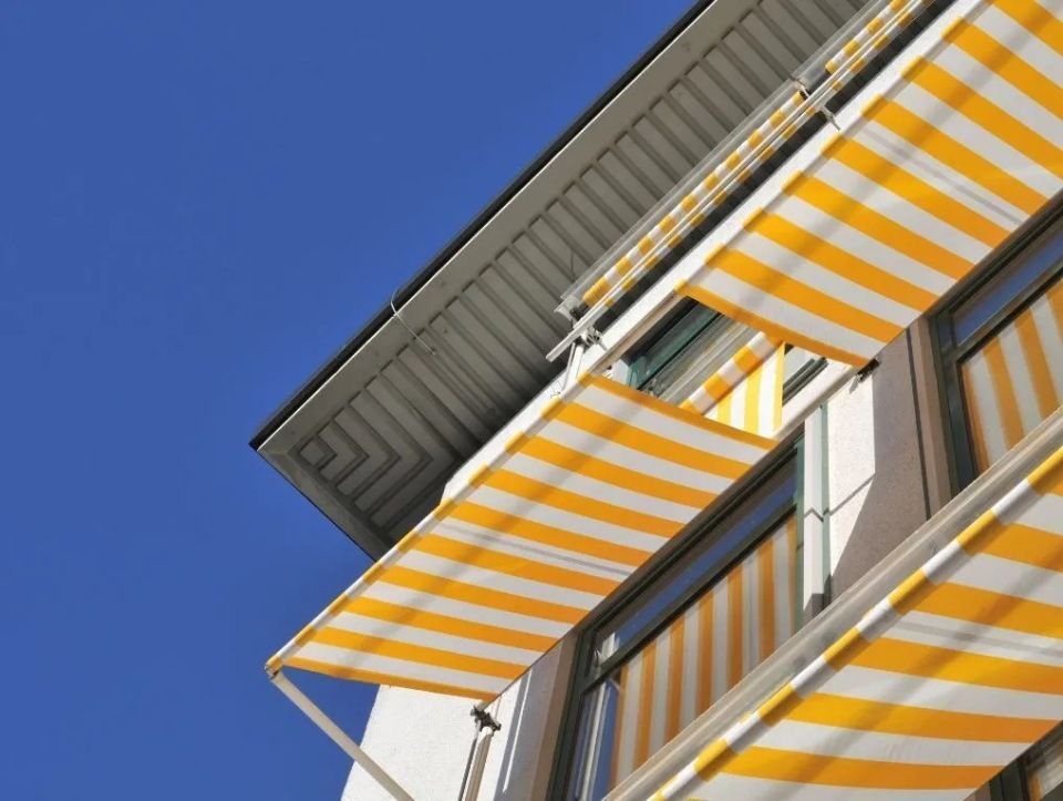 Gelb-weiss gestreifte Markisen spenden den Fenstern eines weissen Gebäudes vor blauem Himmel Schatten.