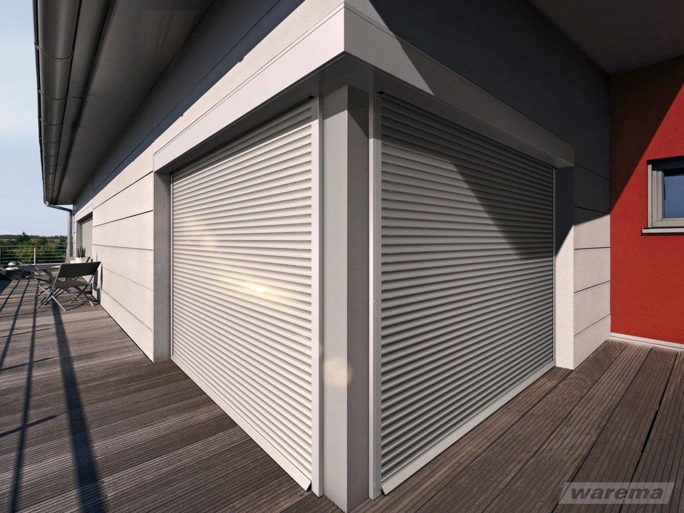 Außenecke eines Gebäudes mit geschlossenen, grauen Rollläden. Holzterrasse und rote Mauer sind sichtbar.