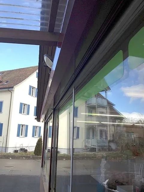 Aussenansicht durch Glastüren, zu sehen ist ein weisses Gebäude mit blauen Fensterläden, die sich im Glas spiegeln.