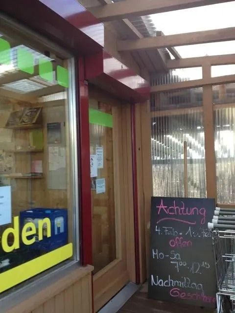 Aussenansicht eines Ladens mit einem Schild. Holztür, Glasfenster und ein Schild mit den Öffnungszeiten in deutscher Sprache.