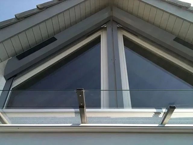 Dreieckiges Fenster mit dunklem Glas, gehalten von Konsolen, unter einem grauen Dach.